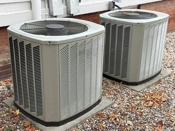 Two outdoor air conditioner condensers sitting on a gravel bed against a brick foundation